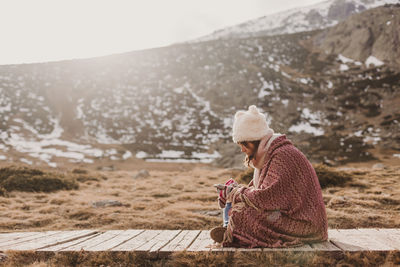 Side view of woman using mobile phone while sitting on footpath against mountain and sky