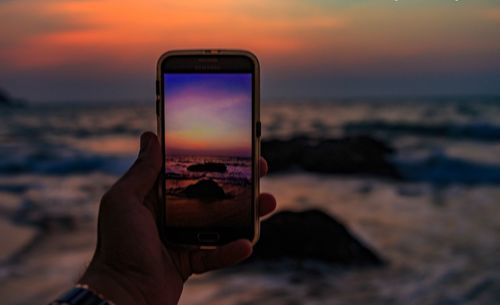 Close-up of hand holding smart phone at beach during sunset
