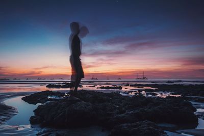 Blur image of silhouette man standing on rock at sea shore against sky during sunset