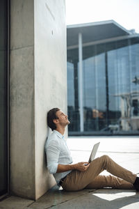 Businessman with laptop looking up sitting by by wall outside building