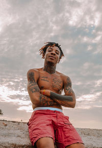 Low angle view of shirtless man with tattoos standing against sky