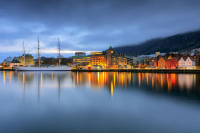 Illuminated buildings reflecting on calm river against cloudy sky