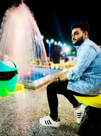 Young man looking away while sitting in illuminated city at night