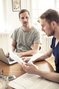 Gay man using laptop while boyfriend reading paper at table in home