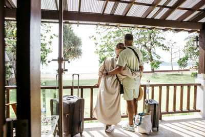 Rear view of male and female tourists with arms around standing on porch of villa