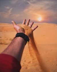 Cropped hand of man falling sand at desert against sky during sunset