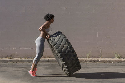 Full length of woman lifting tire truck while exercising against wall