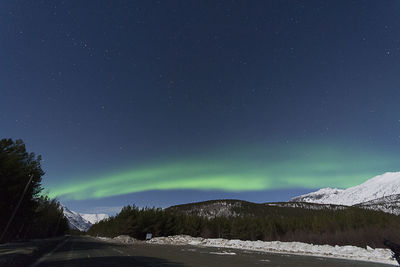 Road by snowcapped mountains against aurora borealis at night