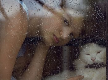 Portrait of young woman with cat at home seen through wet glass