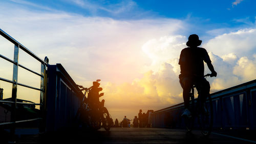 Silhouette man riding bicycle on footbridge against cloudy sky during sunset