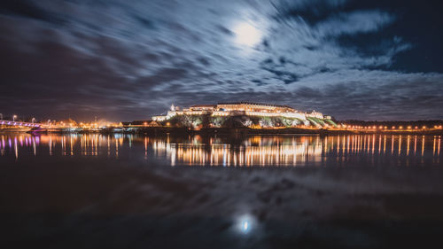 Illuminated buildings by sea against sky at night