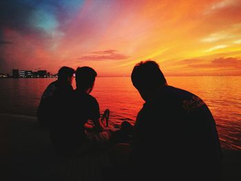 Silhouette friends sitting by sea against sky during sunset