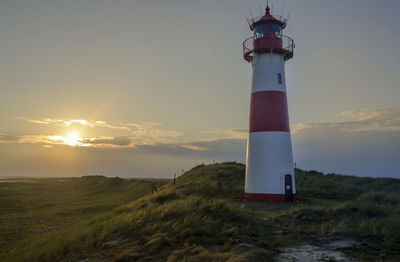 Lighthouse on field by building against sky during sunset