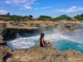 Full length of woman sitting on cliff by sea against sky