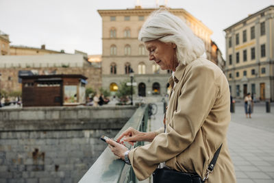 Side view of senior woman leaning on railing while using smart phone in city