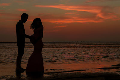 Silhouette friends standing on beach against orange sky