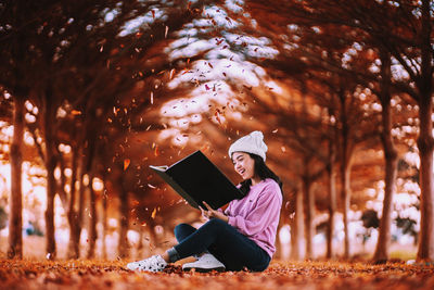 Cheerful young woman reading book while sitting in park during autumn