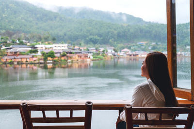 A young woman sitting on balcony and looking at a beautiful lake in mountains village