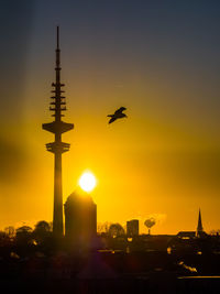 Silhouette bird flying over buildings against sky during sunset