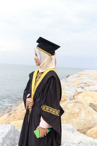 Young woman in graduation gown standing at beach against cloudy sky