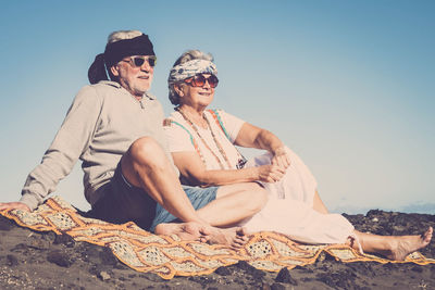 Senior couple wearing sunglasses while sitting at beach against clear sky