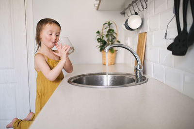 European child of 4 years with water for drink with glass in sink on bright kitchen in real interior