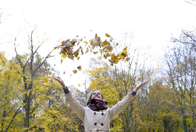 Happy woman throwing leaves while standing against trees during autumn