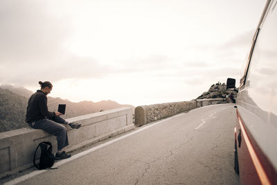 Man using laptop while sitting roadside near van on vacation