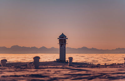 Lighthouse on beach by sea against sky during sunset