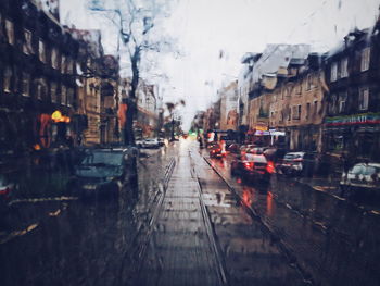 Cars moving on road by buildings in city seen through wet window during rainy season