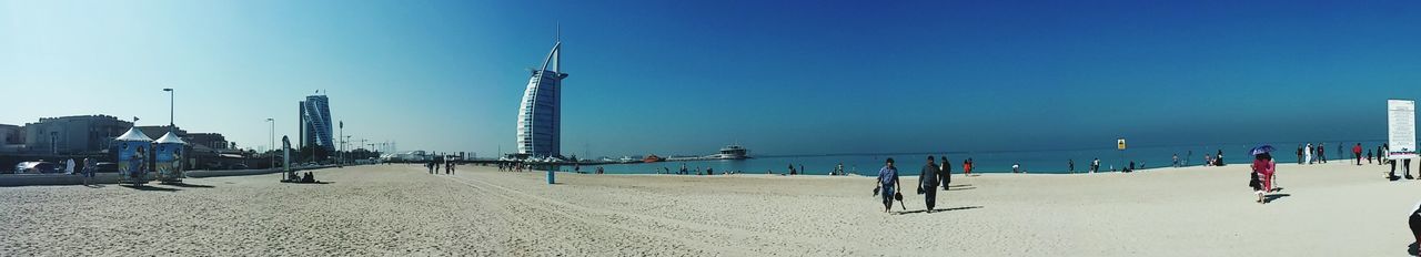 beach, sea, built structure, clear sky, sand, large group of people, water, blue, copy space, architecture, sky, shore, leisure activity, men, building exterior, lifestyles, person, incidental people, day