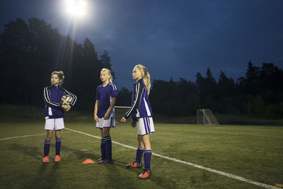 Girls standing on soccer field against sky at dusk
