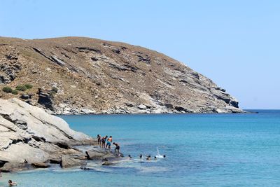 People on rock by sea against clear blue sky