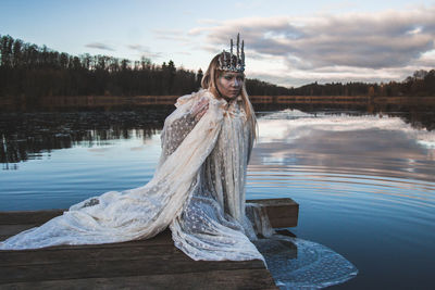 Thoughtful woman wearing crown and white costume sitting on pier by lake during sunset