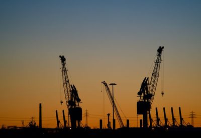 Silhouette cranes at construction site against clear sky during sunset