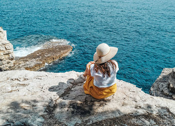 Young woman in yellow skirt sitting on edge of cliff above sea.