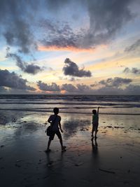 Silhouette boys enjoying at beach against cloudy sky at dusk