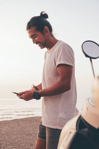 Side view of positive modern sporty male in t shirt and shorts standing next to bike and using mobile phone at seaside at sunset time
