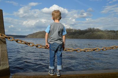 Rear view of boy looking at sea while standing on retaining wall against sky