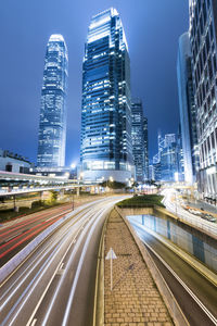 Light trails on city street by buildings against sky at night