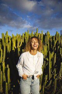 Happy young woman with hands in pockets standing in front of cactus