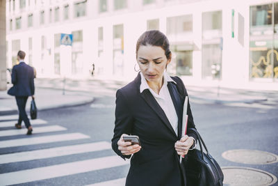 Businesswoman looking at smart phone while crossing road in city