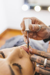 Cheerful blond woman with closed eyes while tattooed makeup artist smearing liquid lipstick on lips of model