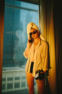 Side view of young female traveler in white shirt and sunglasses with towel wrapped around head talking on telephone in modern hotel room