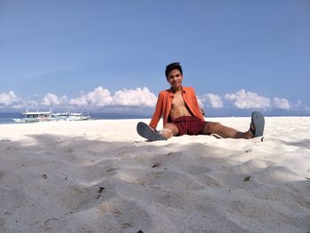 Portrait of young man sitting on sand at beach against sky