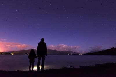 Rear view of father and daughter standing on lakeshore against sky at night