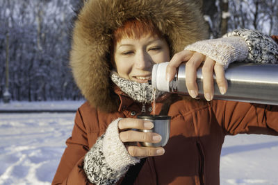 Smiling woman in red coat drinks hot tea from metal thermos. leisure activity in winter forest