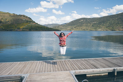 Young woman jumping on pier over lake against sky