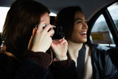 Woman photographing through camera while sitting with happy friend in taxi