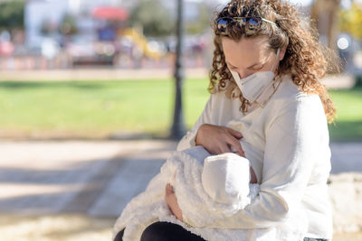 Mother wearing mask breastfeeding baby while sitting outdoors